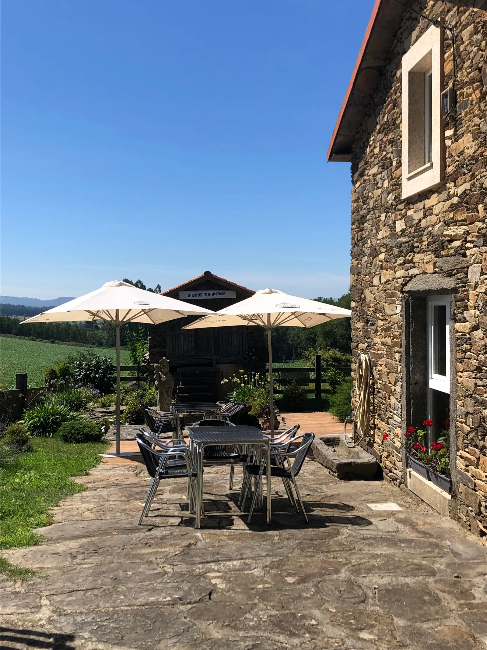Steinhaus mit Terrasse, Sonnenschirmen und Bestuhlung, dahinter grüne Hügellandschaft unter blauem Himmel