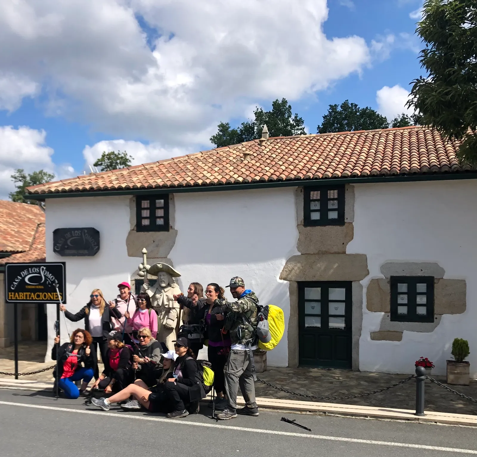Pilgergruppe singt fröhlich vor weißem Steinhaus mit Ziegeldach, Fenster und Tür in dunklem Holz, blauer Himmel mit Wolken