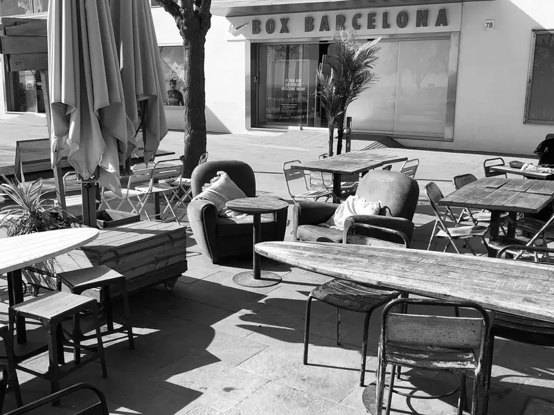 Leere Terrassencafé mit Tischen und Stühlen, im Hintergrund das Schild "Box Barcelona", Sonnenschatten auf dem Boden