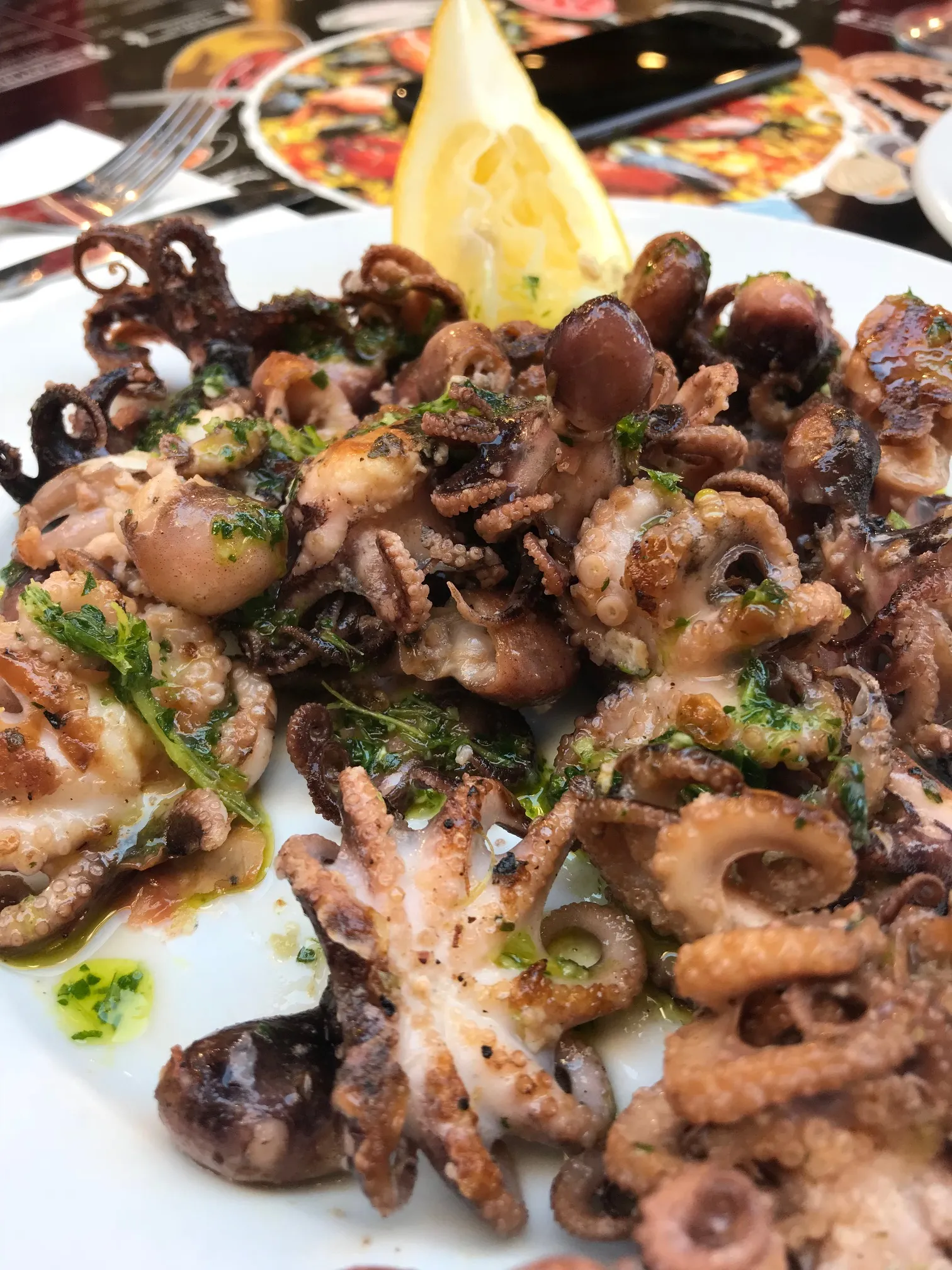 Gegrillter Oktopus mit Zitrone und Petersilie auf weißem Teller, garniert mit Öl, vor unscharfem Restaurant-Hintergrund