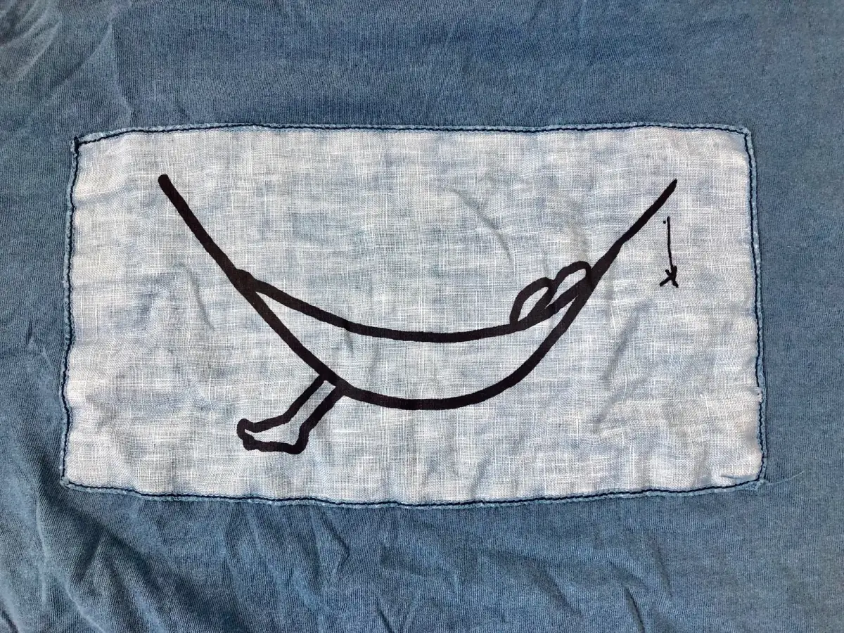 Blaugraues Shirt mit aufgestickter schwarzer Hängematte auf heller Leinenfläche, umrahmt von dunklem Stoff