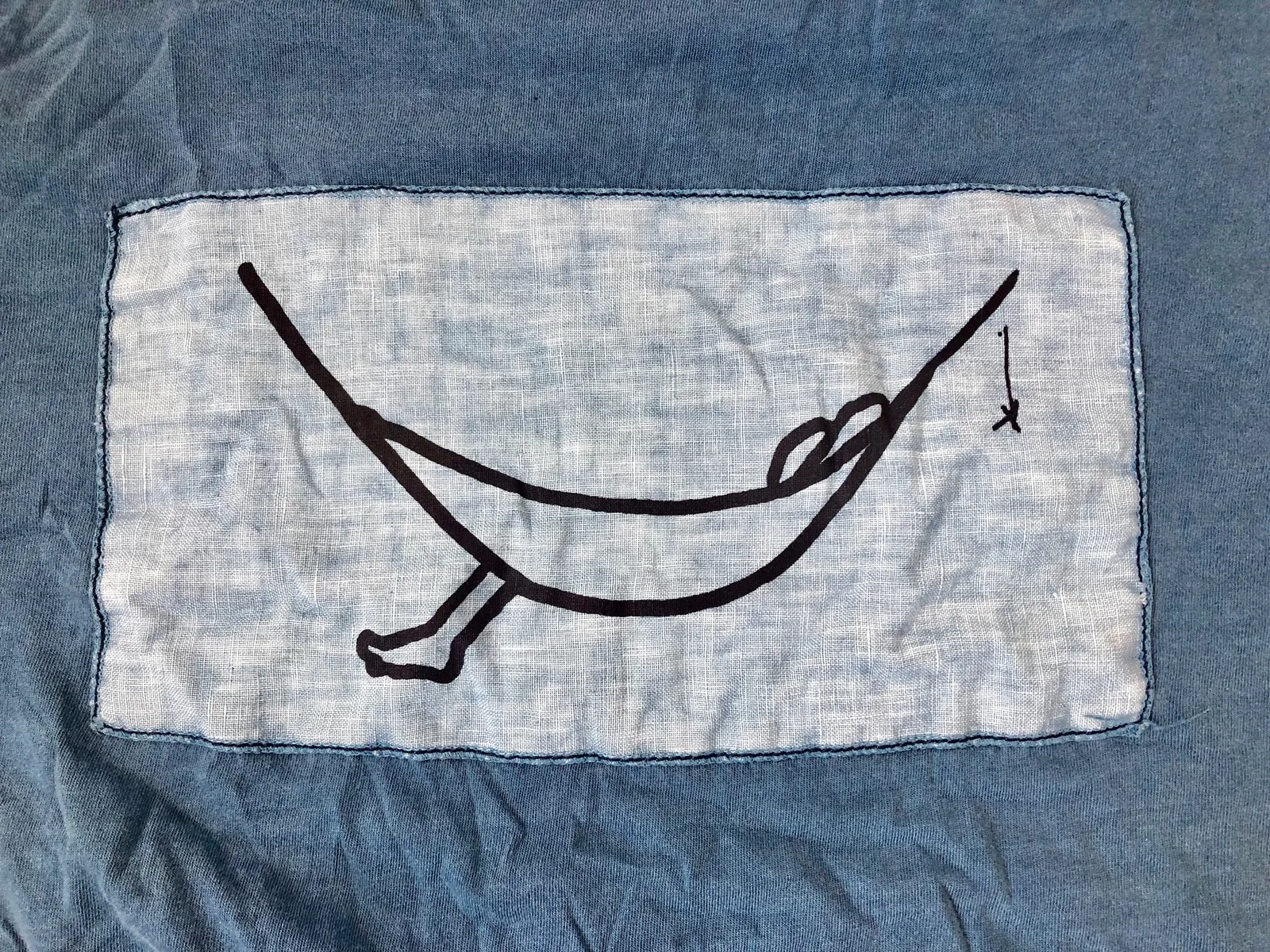 Blaugraues Shirt mit aufgestickter schwarzer Hängematte auf heller Leinenfläche, umrahmt von dunklem Stoff