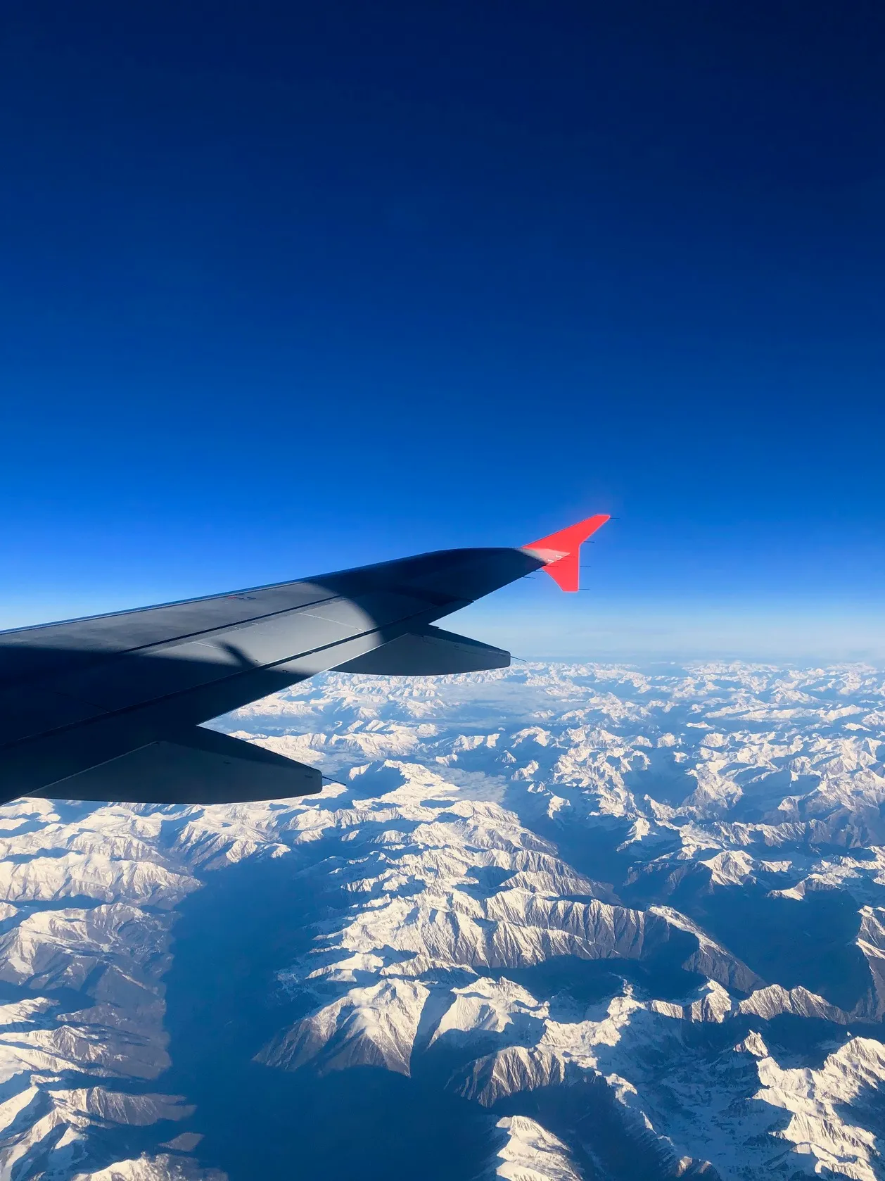 Flugzeugflügel mit rotem Winglet über schneebedeckten Alpen unter stahlblauem Himmel