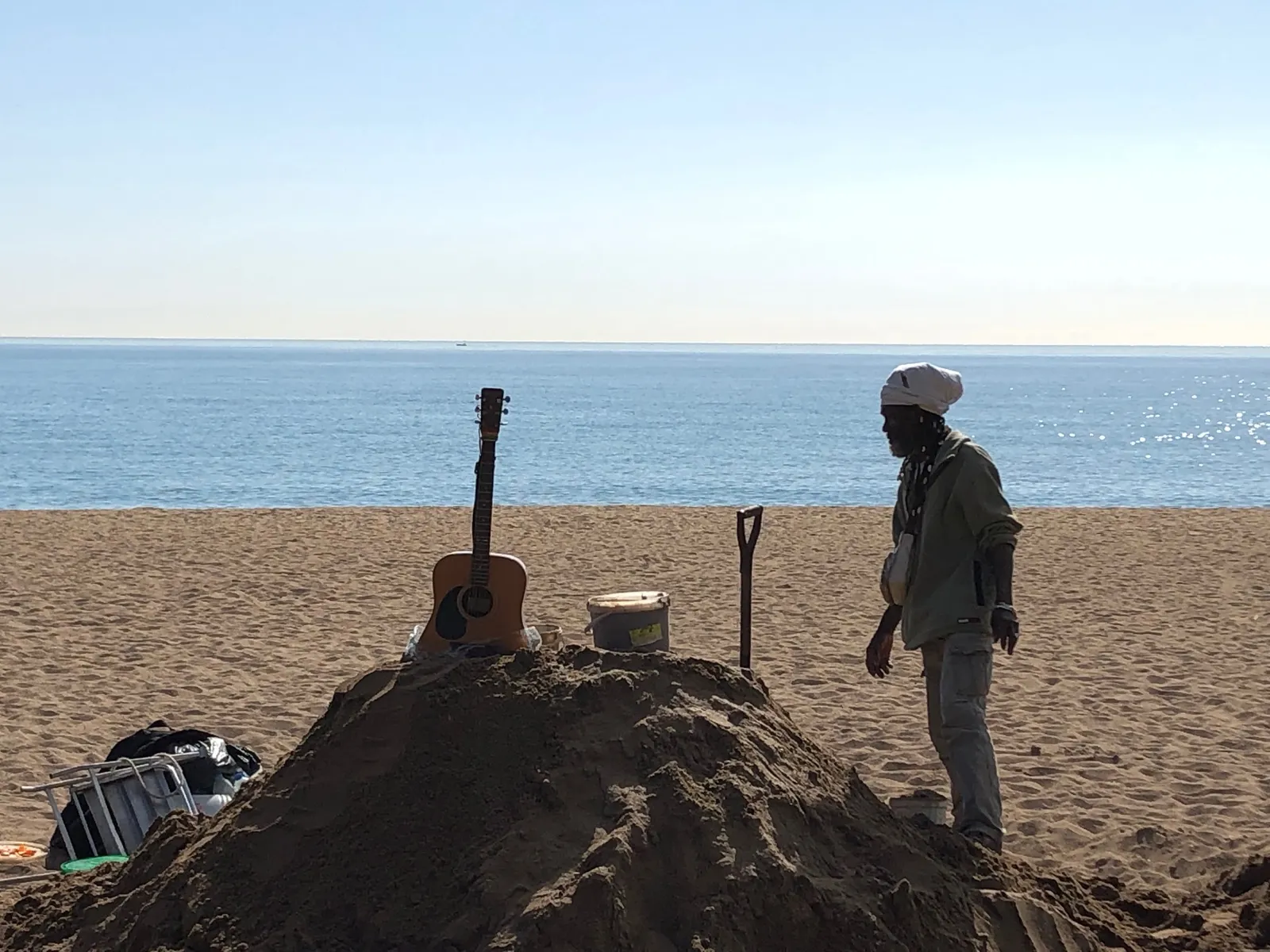 Mann mit Kappe steht neben Sandskulptur mit Gitarre, Spatenund Behälter am Strand, Meer im Hintergrund
