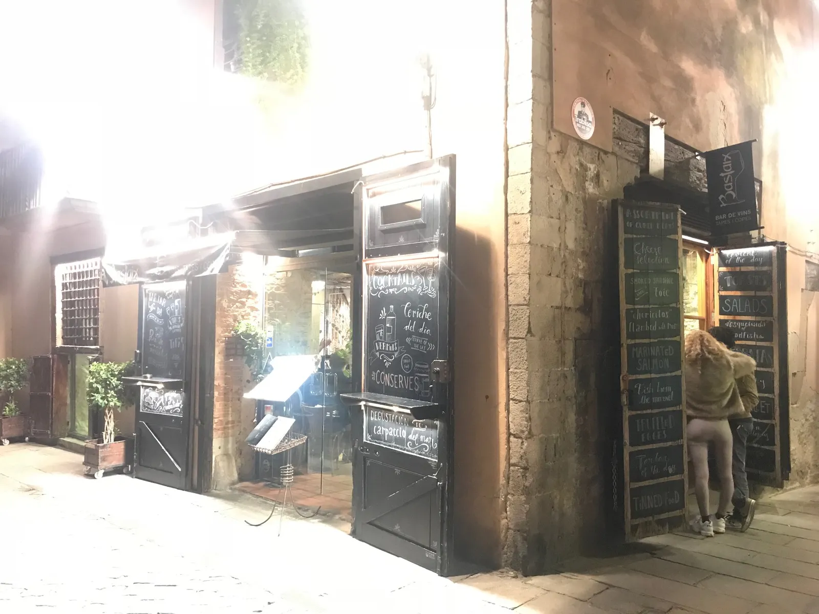 Schmale Gasse mit kleinen Restaurants und Bars, schwarze Türen mit Kreidetafeln, Person vor Eingang, warmes Abendlicht