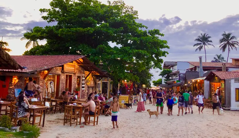 Strandpromenade bei Sonnenuntergang mit lebhaftem Treiben, Menschen flanieren zwischen beleuchteten Restaurants und Häusern