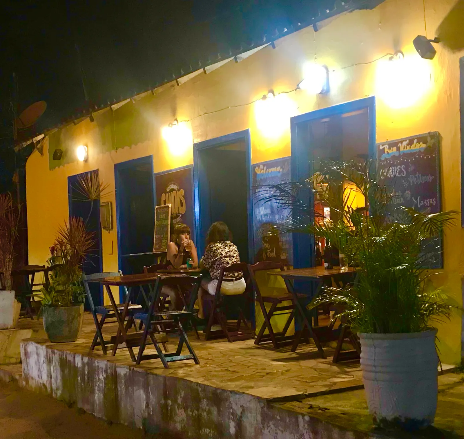 Abendliche Strandbar mit gelbem Gebäude und blauen Fenstern, Gäste sitzen an Tischen, Livemusik-Atmosphäre unter Lampen