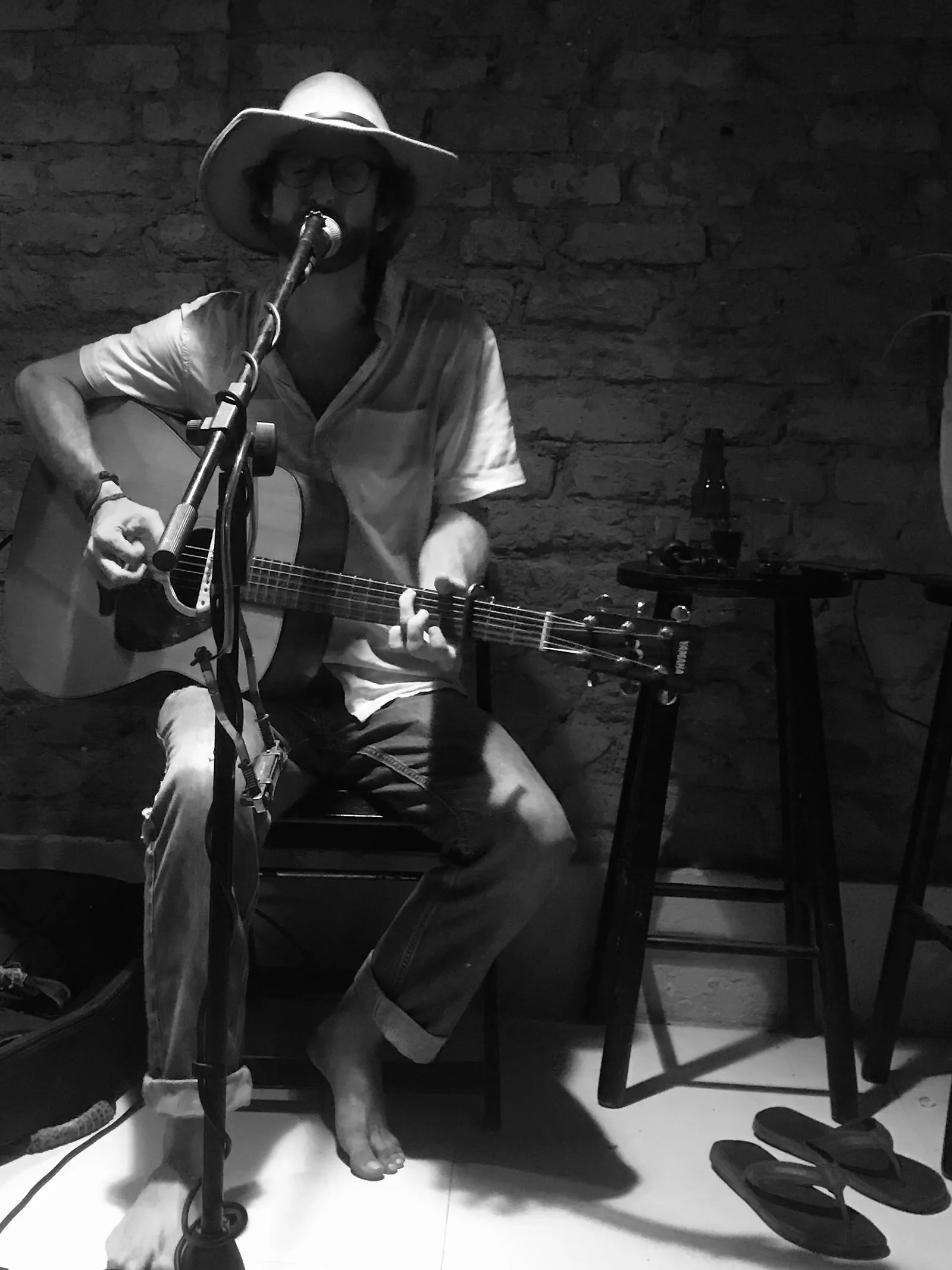 Amerikanischer Sänger sitzt auf Hocker mit Gitarre vor Backsteinmauer, trägt Cowboyhut und singt ins Mikrofon, barfuß