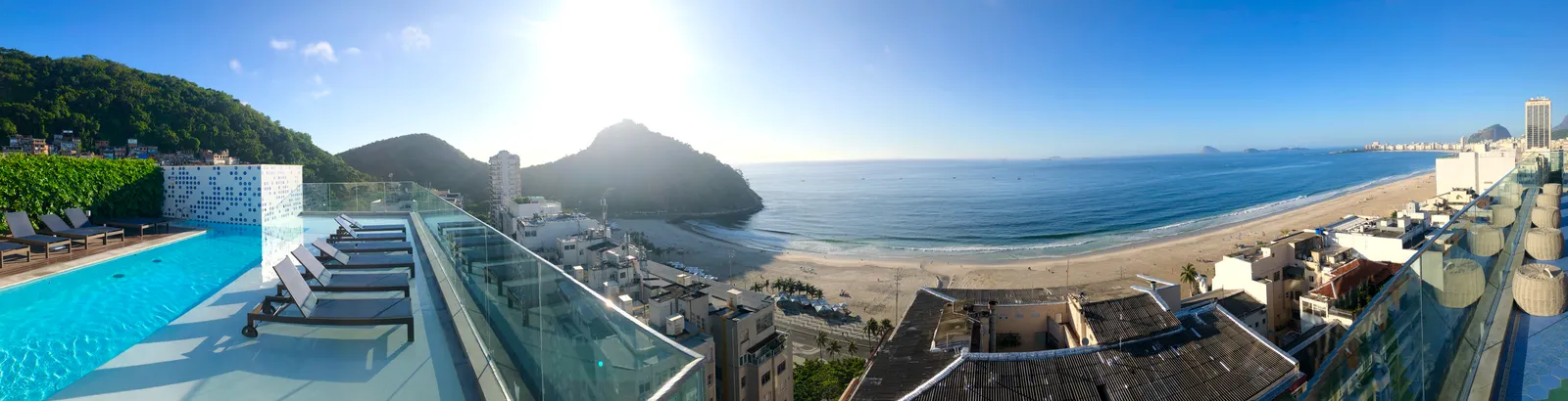 Panoramablick auf Strand und Bucht mit Pool im Vordergrund, grünen Bergen und blauem Meer unter sonnigem Himmel