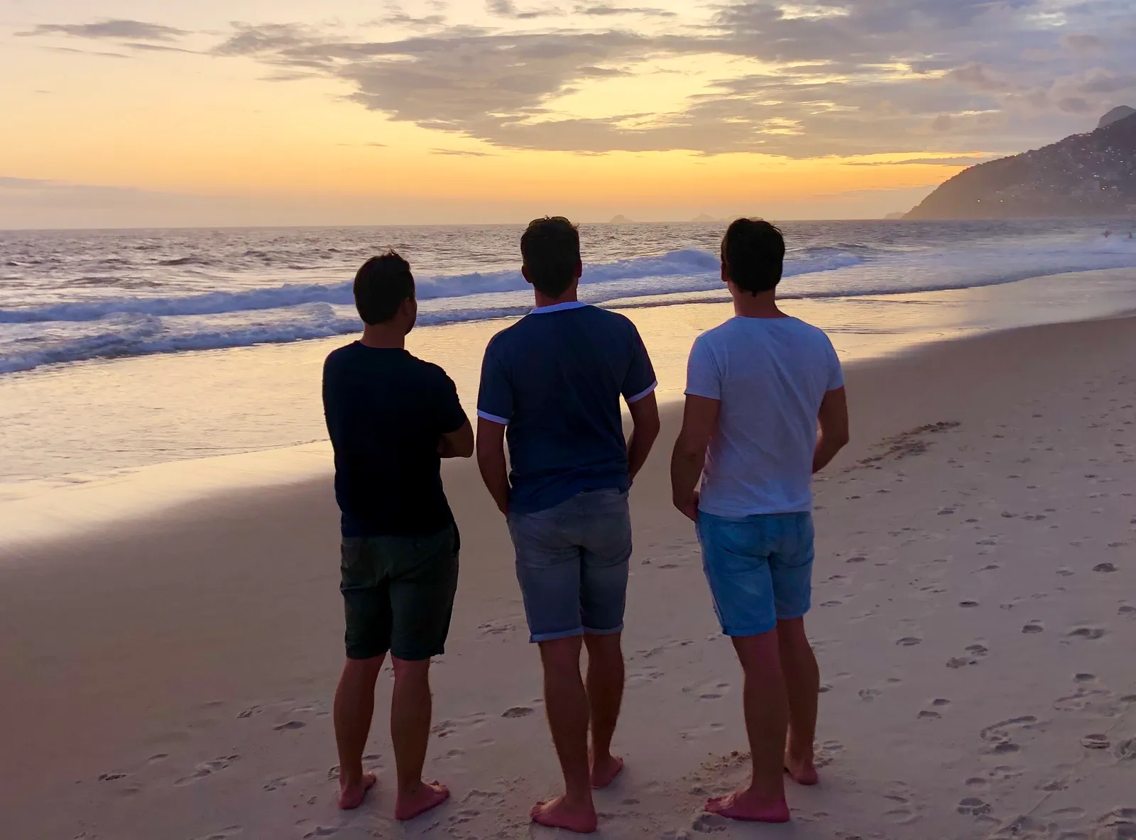 Drei Männer stehen bei Sonnenuntergang am Strand und blicken auf das Meer mit Wellen und einer Bergkulisse