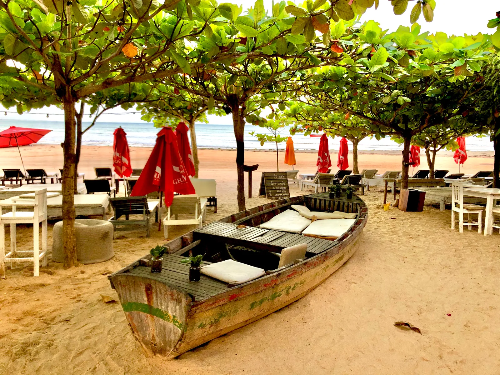 Strandbar unter Laubdach mit altem Fischerboot als Sitzmöbel, roten Schirmen und Liegestühlen am Sandstrand