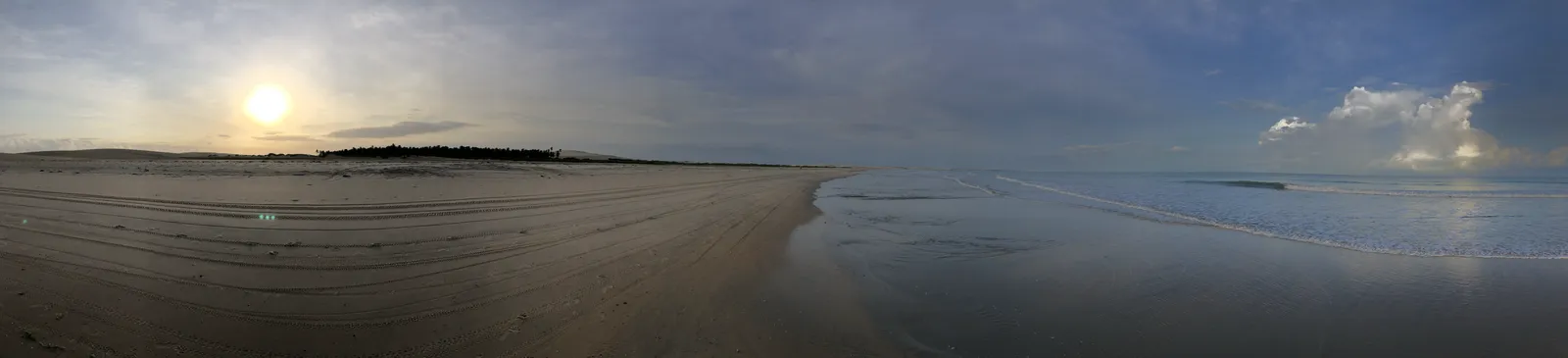 Breiter Sandstrand bei Sonnenuntergang mit ruhiger Wasserfläche und Dünen unter bewölktem Himmel