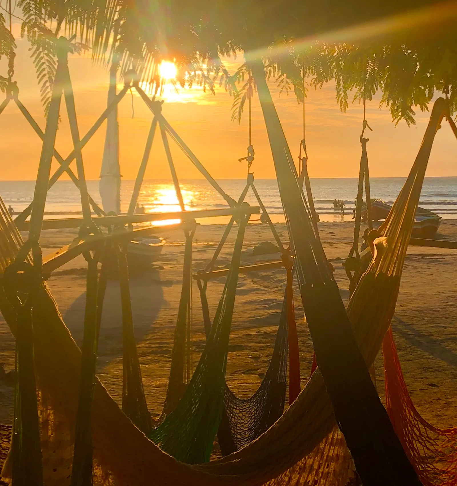 Sonnenuntergang am Strand mit Hängematten zwischen Palmen und Holzpfählen, entspannte Strandatmosphäre in warmen Goldtönen