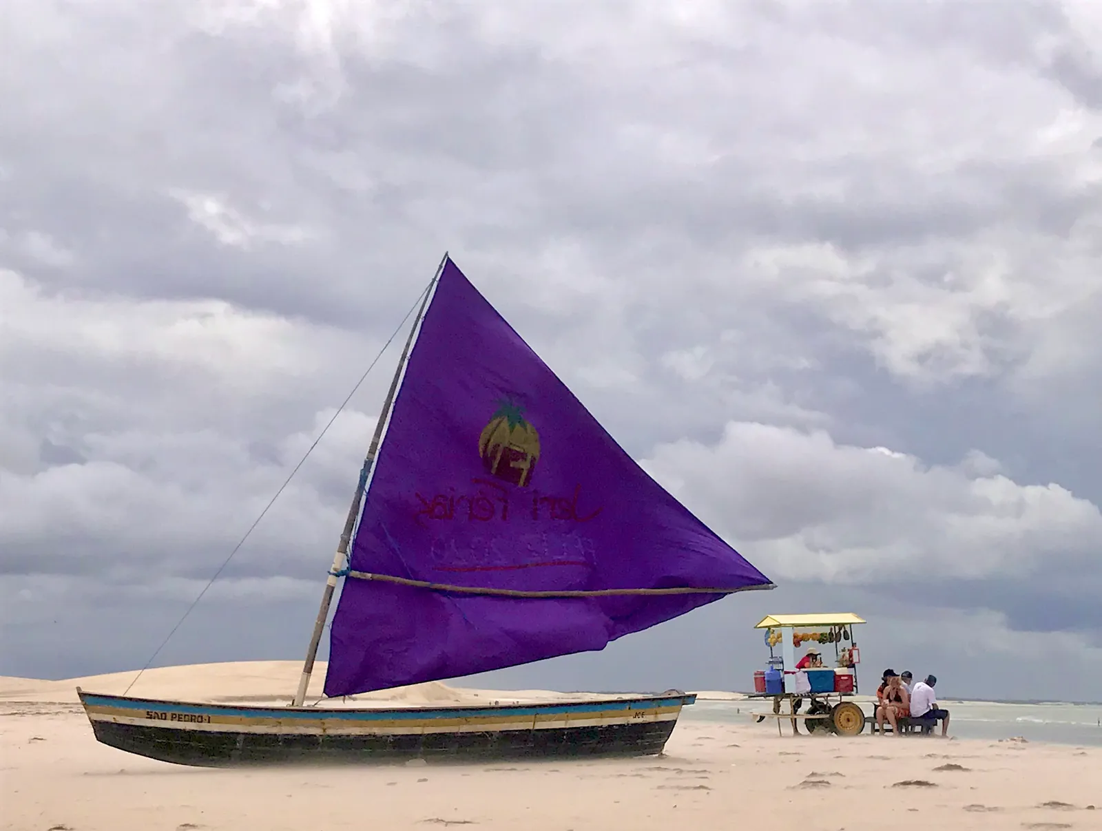 Fischerboot mit violettem Segel am Strand, im Hintergrund mobile Verkaufsstände und Menschen unter bewölktem Himmel