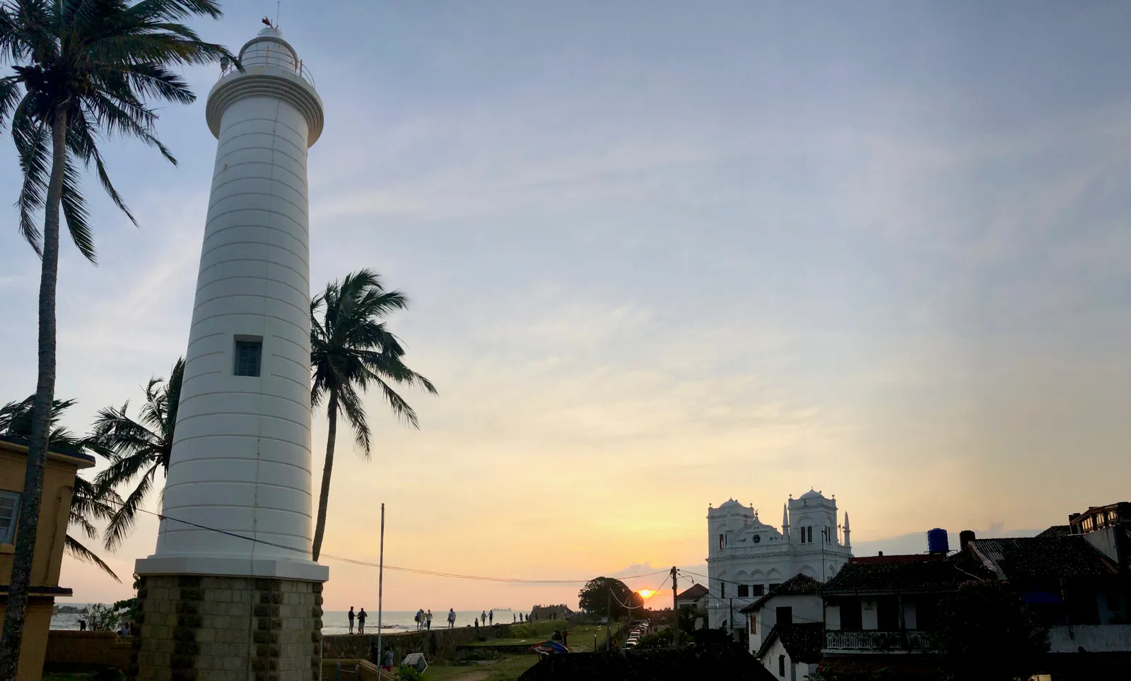 Weißer Leuchtturm bei Sonnenuntergang mit Palmen, historischen Gebäuden und Menschen auf der Festungsmauer von Galle
