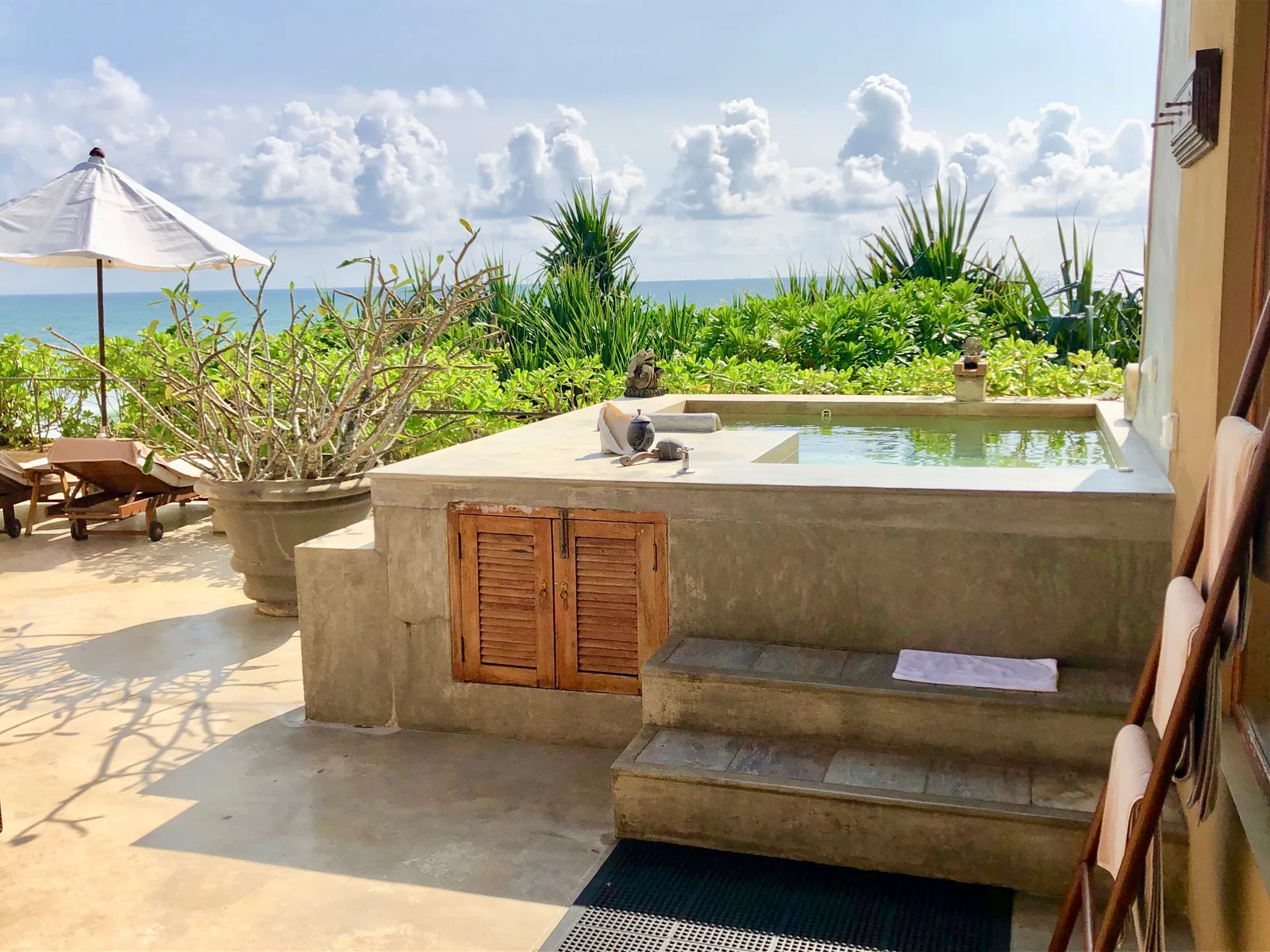 Whirlpool auf einer Terrasse mit Meerblick, umgeben von tropischen Pflanzen, Sonnenschirm und Liegestühlen an der Westküste
