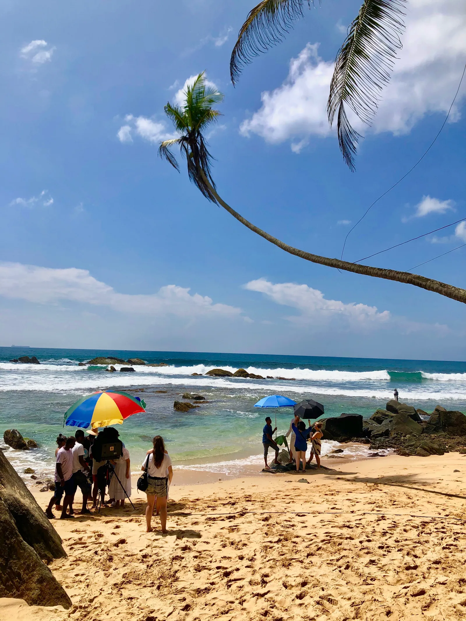 Strand in der Bucht von Dalawella mit Besuchern unter bunten Sonnenschirmen, im Hintergrund Wellen und eine gebogene Palme