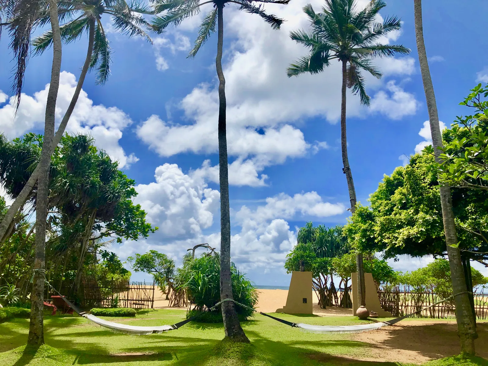 Tropische Gartenanlage mit Palmen, Rasenflächem und traditionellen Strukturen an einem Strand unter blauem Himmel