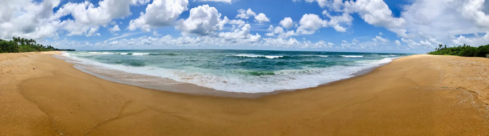 Breiter Sandstrand mit Wellen, die auf den Strand zulaufen, türkises Meer und bewölkter blauer Himmel
