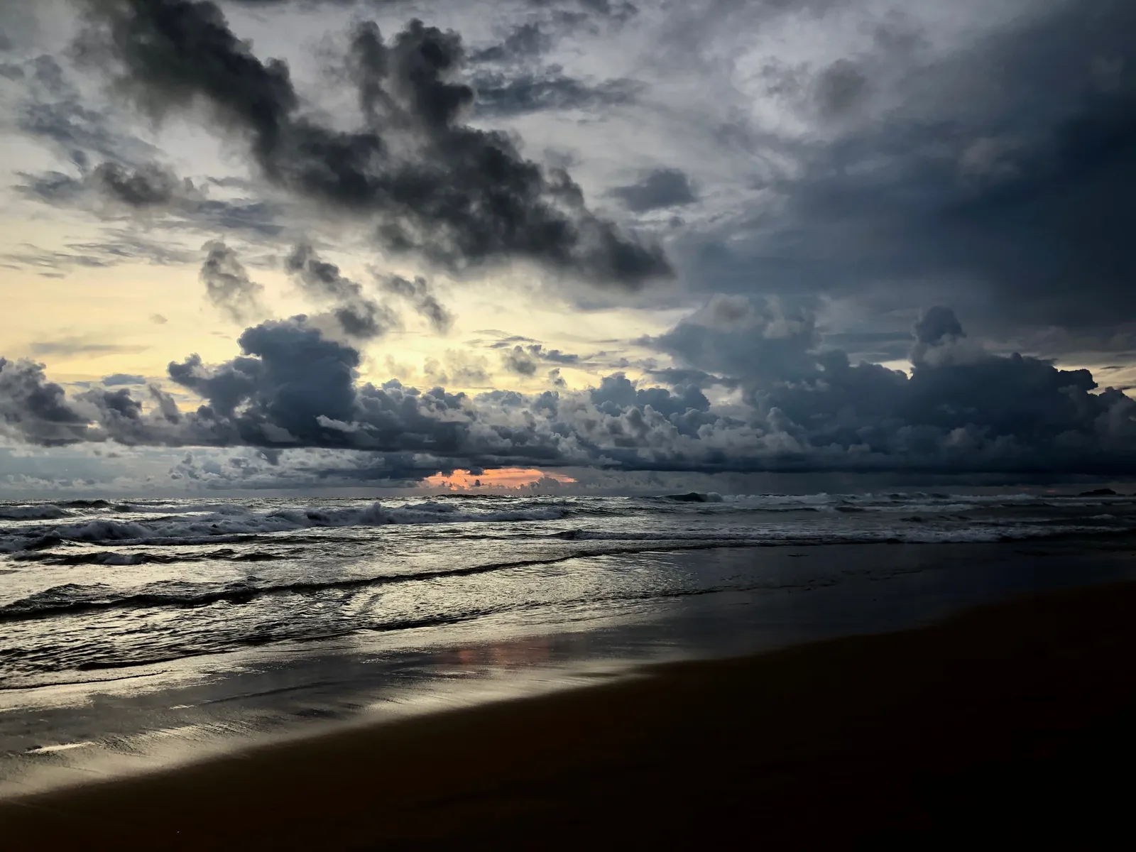 Strand bei Sonnenuntergang mit dramatischem Wolkenhimmel, orange leuchtende Sonne über dem Meer, Wellen am Ufer