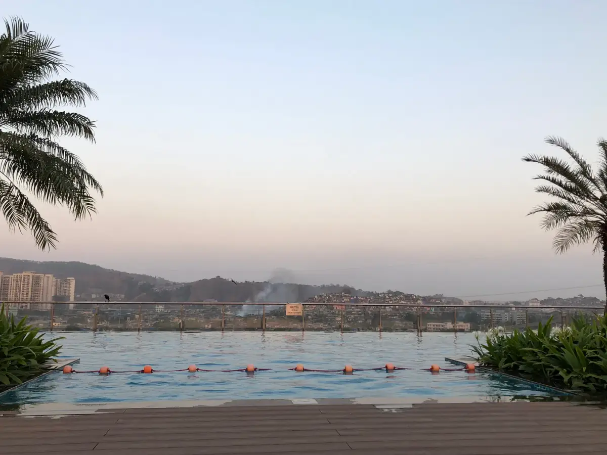 Infinity-Pool mit orangefarbenen Bojen vor Flusslandschaft und Hochhäusern in diesiger Morgenstimmung, Palmen im Vordergrund