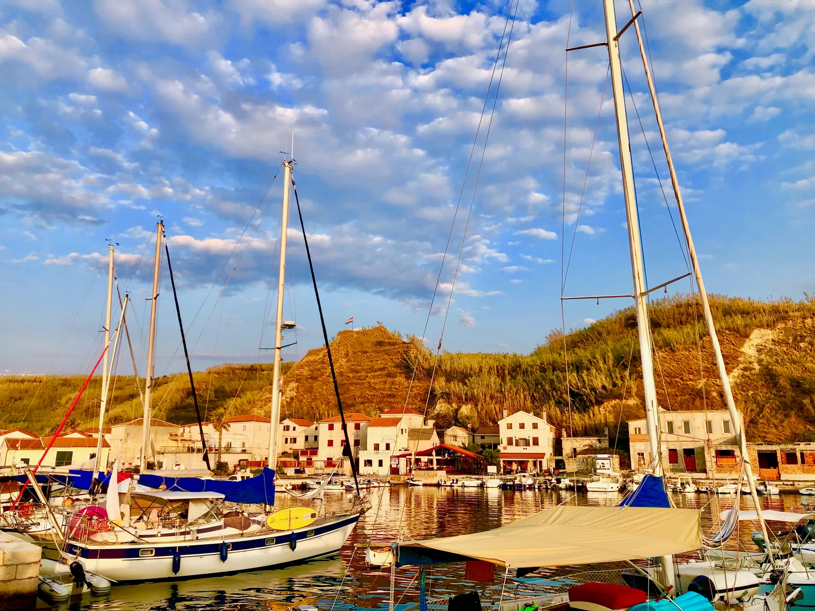 Segelboote mit weißen Masten liegen in einem idyllischen Hafenbecken, dahinter Häuser am Hang unter blauem Himmel mit Wolken