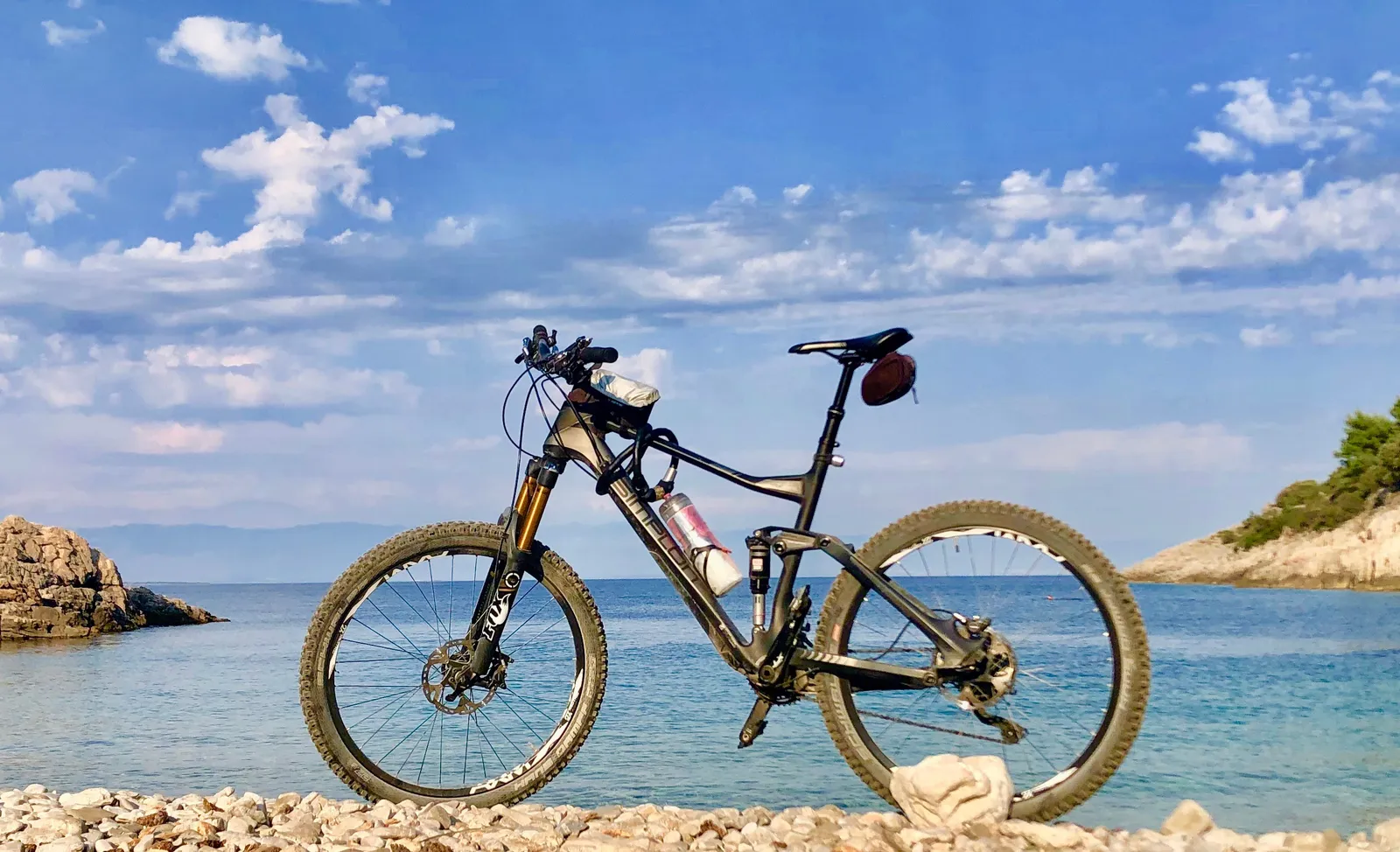 Mountainbike steht auf Kieselstrand vor türkisfarbenem Meer und felsiger Küstenlandschaft unter blauem Himmel
