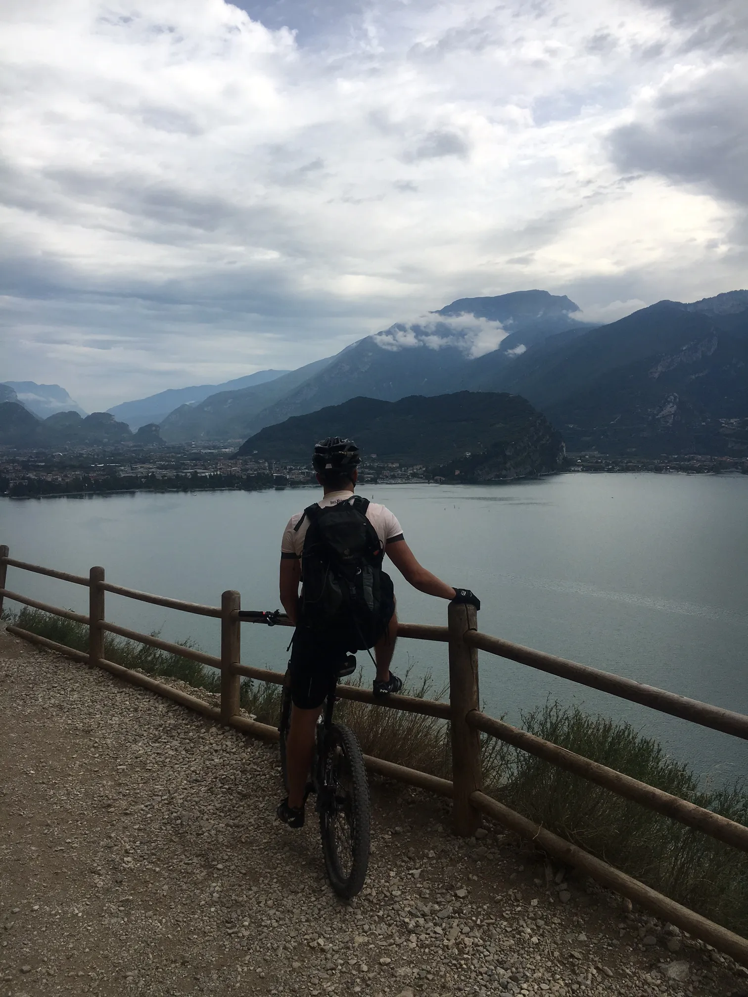 Mountainbiker auf Waldweg mit Holzgeländer blickt auf Bergsee und bewaldete Bergketten unter Wolkenhimmel