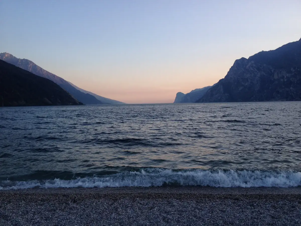 Alpensee bei Sonnenuntergang mit Steilküsten, ruhige Wasserfläche und kleine Wellen am Kiesstrand in blauer Dämmerung