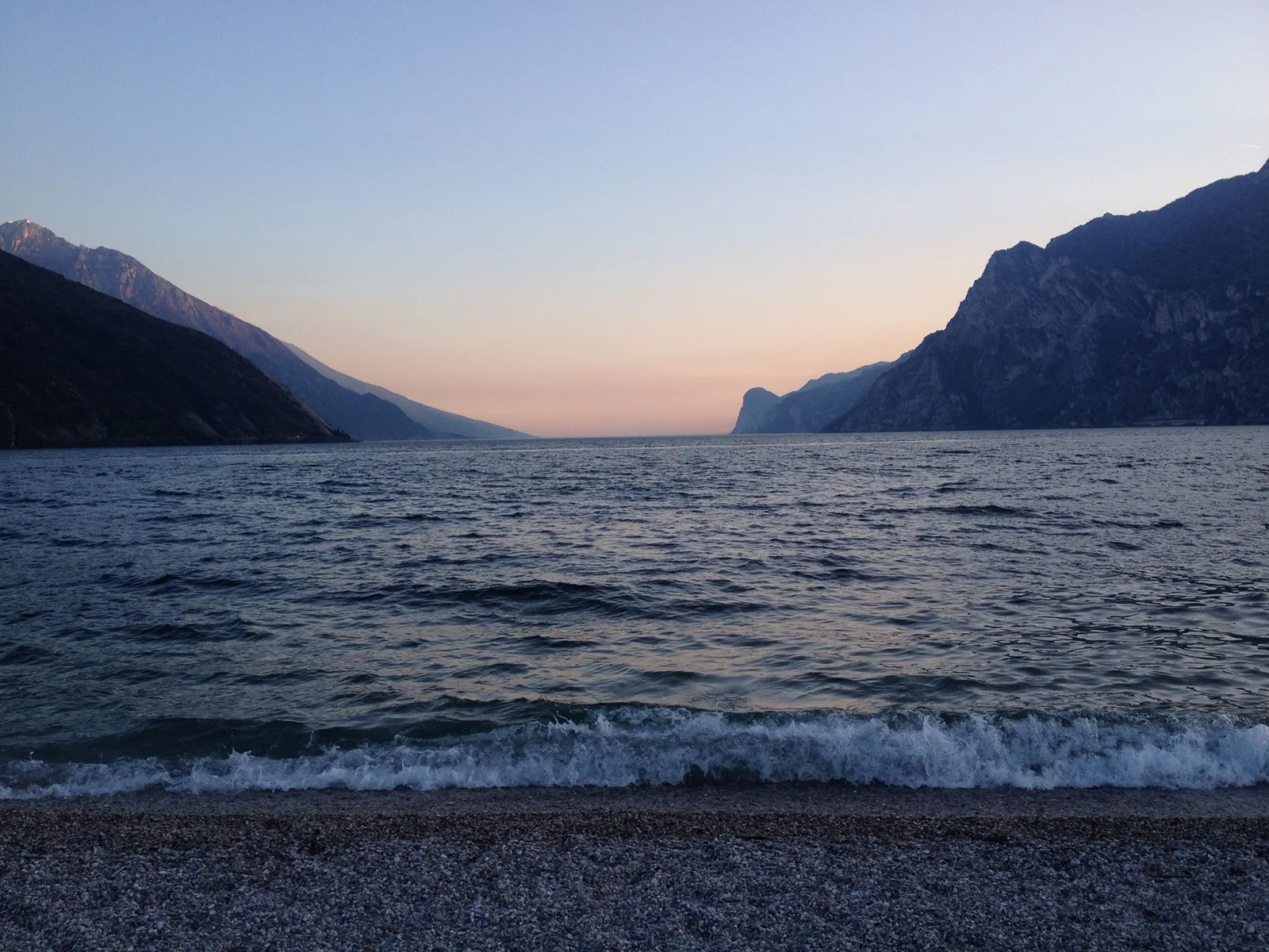 Alpensee bei Sonnenuntergang mit Steilküsten, ruhige Wasserfläche und kleine Wellen am Kiesstrand in blauer Dämmerung