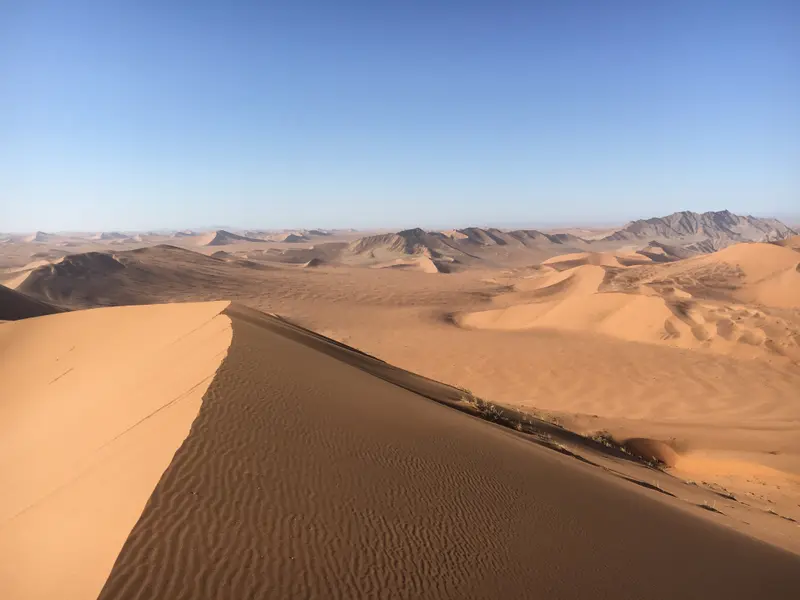Schotterband durchzieht endlose Wüstenlandschaft mit Sanddünen und fernen Bergen unter klarem Himmel