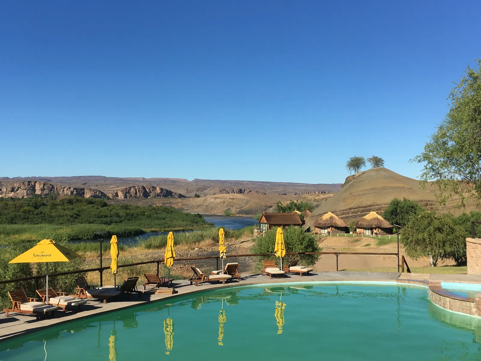 Luxuriöser Pool mit gelben Sonnenschirmen vor dramatischer Berglandschaft und traditionellen Lehmhütten am Fluss in Namibia
