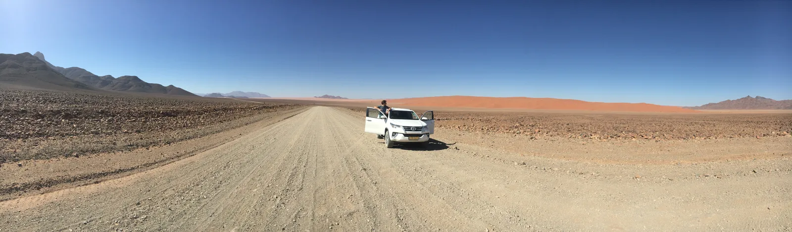 Weißer Jeep auf endloser Schotterstraße durch karge Wüstenlandschaft unter blauem Himmel