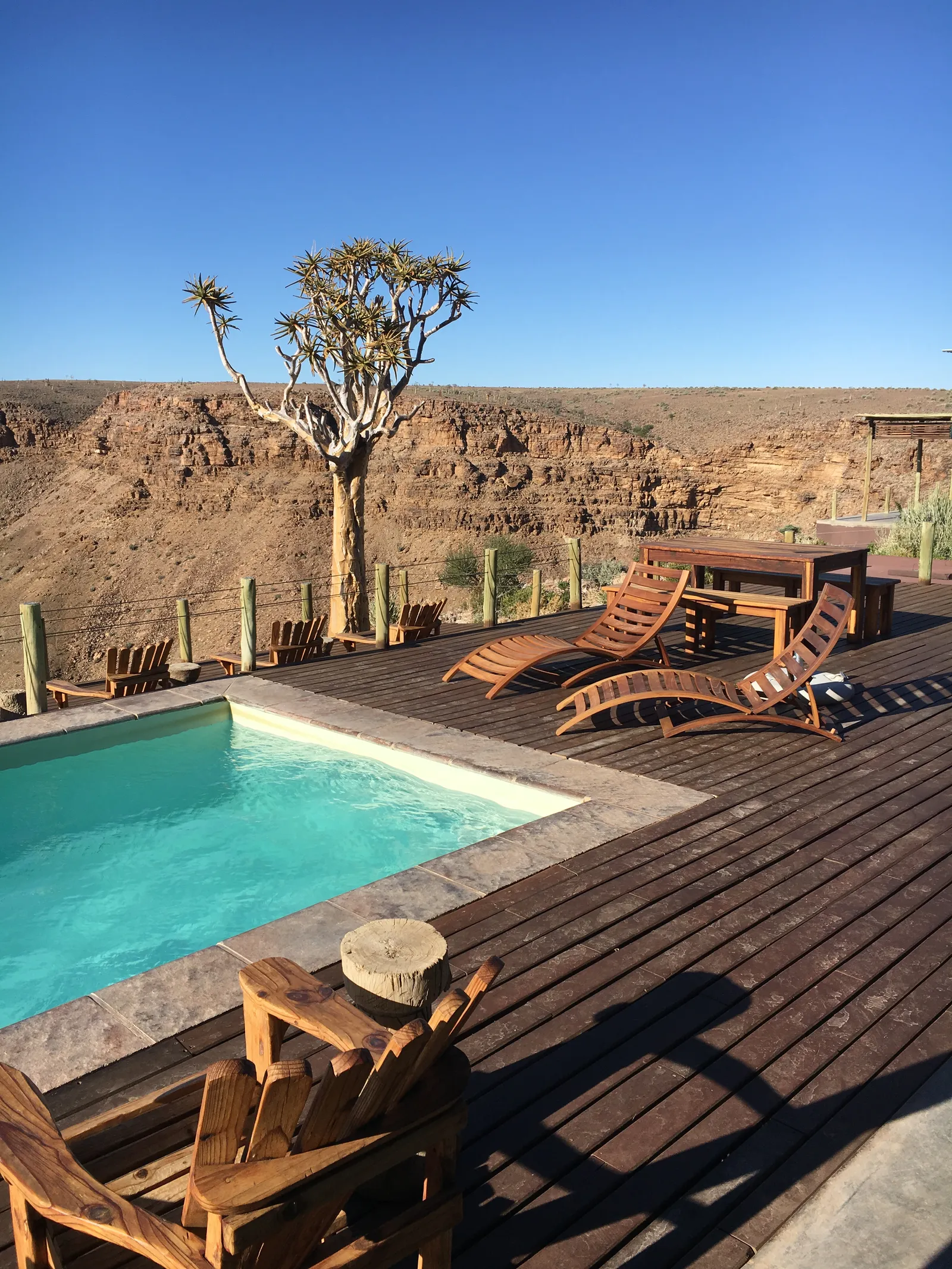 Luxuriöse Poolanlage mit Holzliegen auf einer Terrasse, dahinter karge Wüstenlandschaft