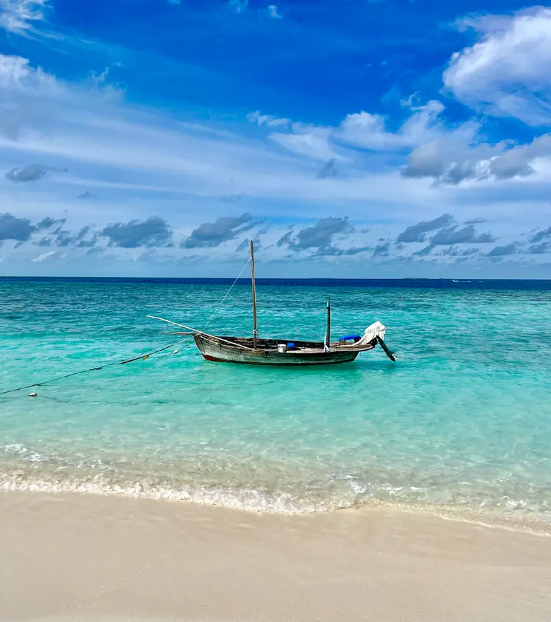 Holzboot mit zwei Masten ankert in türkisblauem Wasser vor Sandstrand unter blauem Himmel mit Cumulus-Wolken