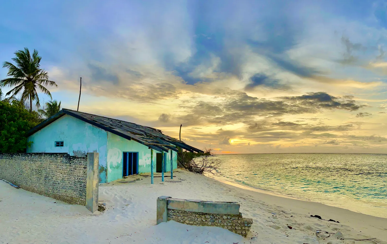 Türkises Strandhaus bei Sonnenuntergang mit Palmen, Steinmauer und ruhigem Meer an abgelegener Küste
