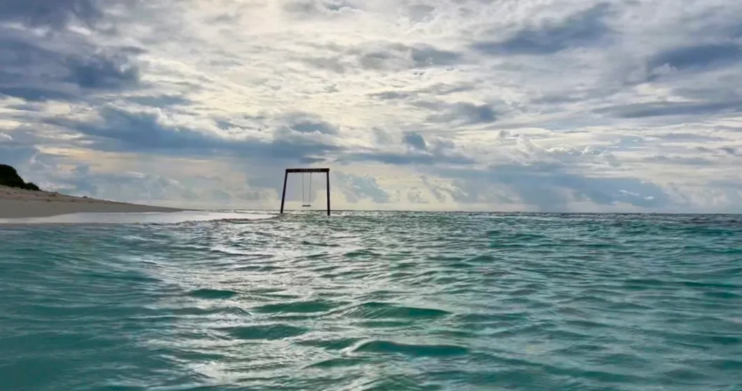 Türkisblaues Meer mit verlassenem Schaukelgerüst im Wasser, bewölkter Himmel und Inselküste im Hintergrund