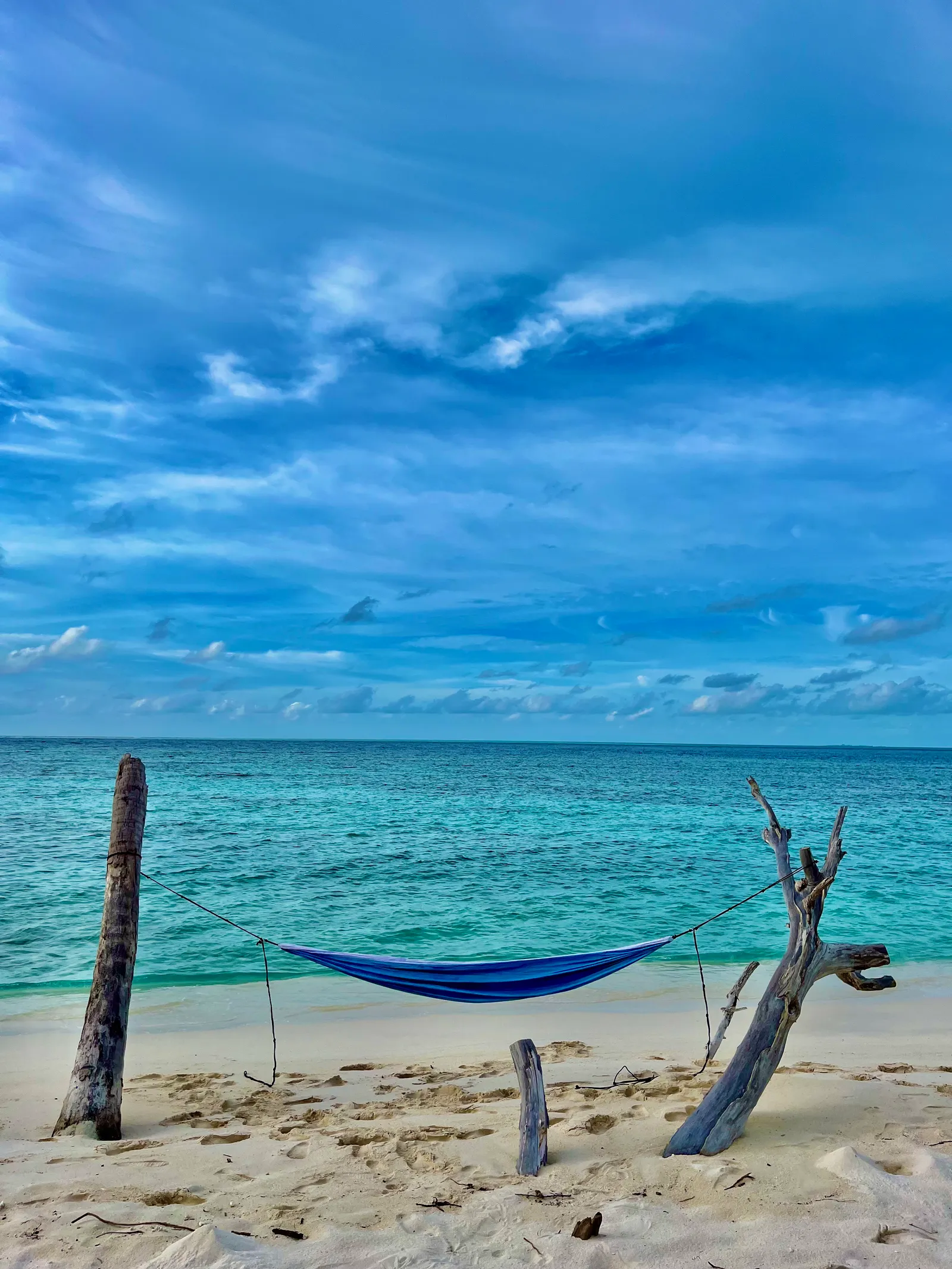 Blaue Hängematte zwischen Treibholzpfählen am Sandstrand, türkisblaues Meer und bewölkter Himmel im Hintergrund
