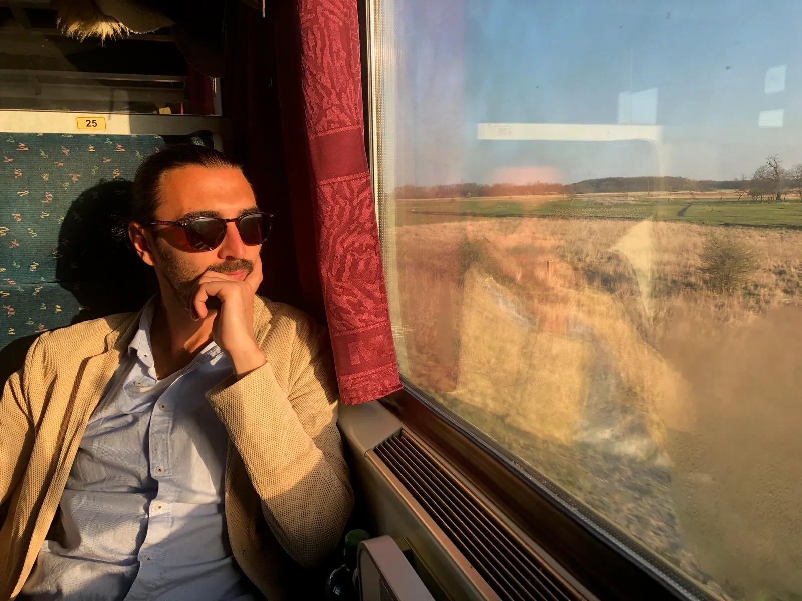Mann mit Sonnenbrille und Schnurrbart blickt nachdenklich aus Zugfenster auf flache Landschaft mit Feldern und Baumreihen