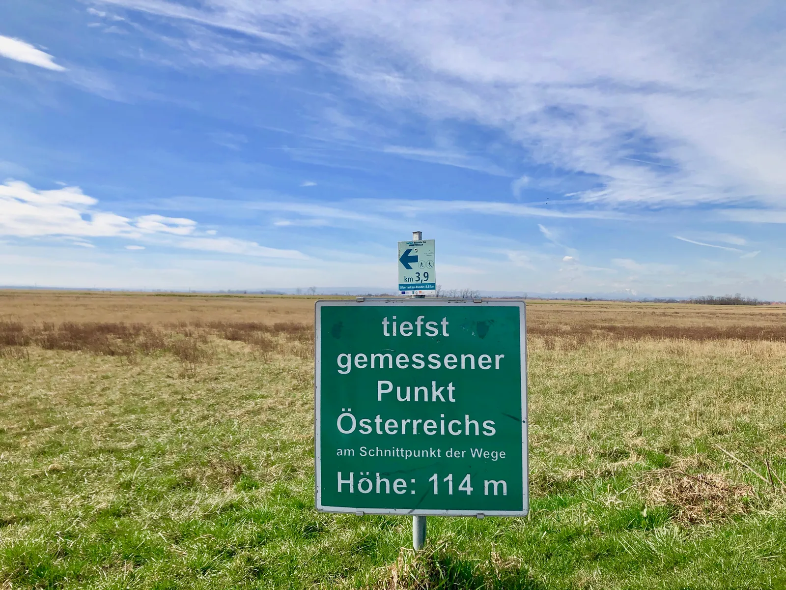 Grünes Schild „tiefst gemessener Punkt Österreichs, Höhe: 114 m" an Feldwegkreuzung in flacher Pannonischer Tiefebene unter