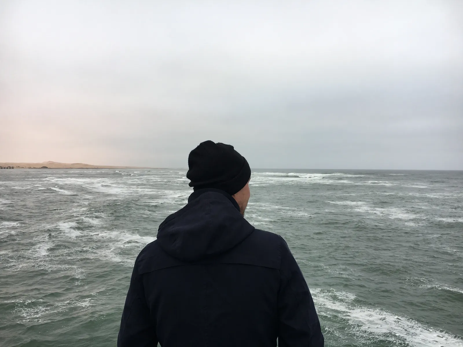 Person in schwarzer Jacke und Mütze steht am Strand von Swakopmund und blickt auf das Meer mit Wellen und grauem Himmel