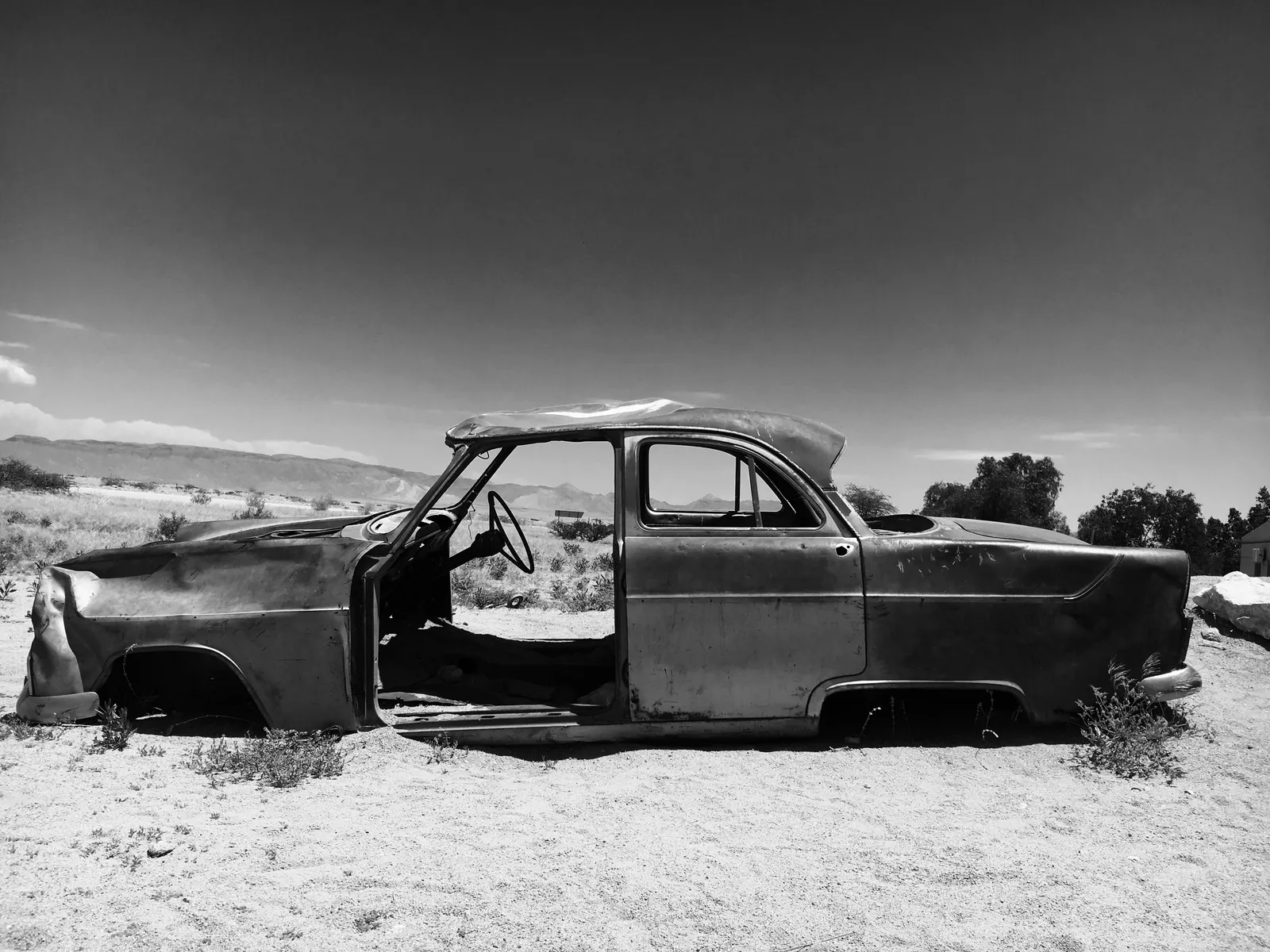 Verlassenes Autowrack mit offener Türe in karger Wüstenlandschaft unter klarem Himmel, verrostet und von Vegetation umgeben