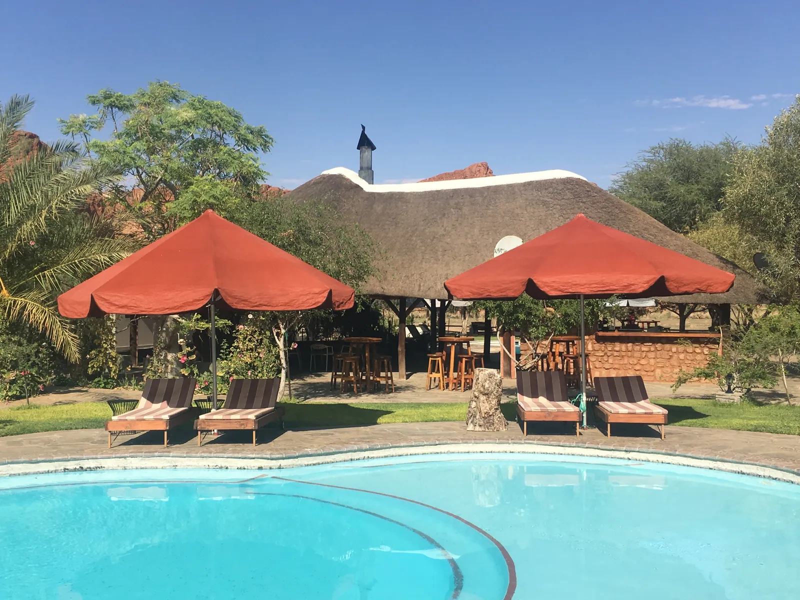Swimmingpool mit türkisblauem Wasser vor einer Lodge mit roten Sonnenschirmen, Liegestühlen und Strohdachhütten in Namibia