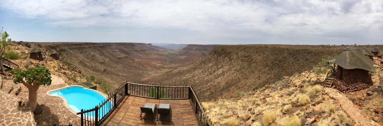 Holzterrasse mit türkisblauem Pool auf Felskante über weitem, trockenem Canyon unter bewölktem Himmel