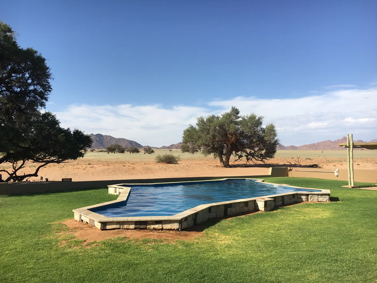 Swimmingpool mit tiefblauem Wasser in einer Grünfläche vor weiter Steppenlandschaft mit Akazienbäumen und fernen Bergen