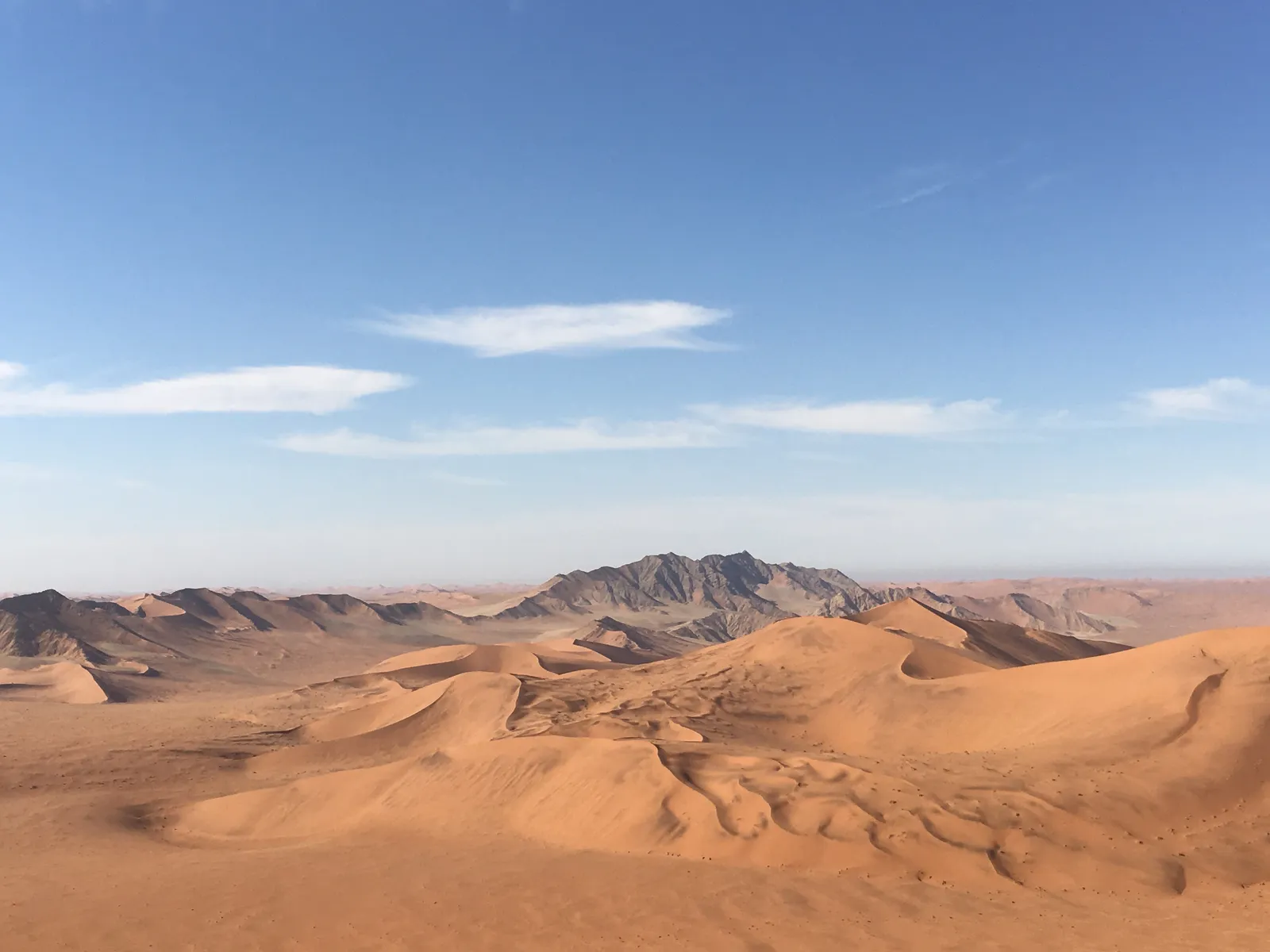 Weite Wüstenlandschaft mit Sanddünen und felsigen Bergen unter blauem Himmel mit leichten Wolken