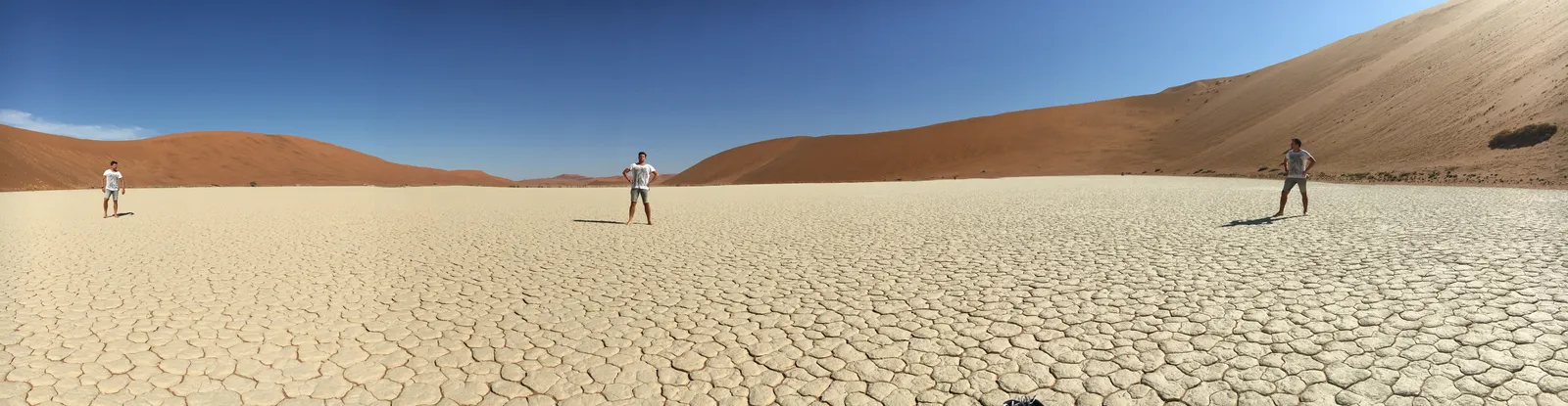 Drei Menschen stehen auf ausgetrocknetem, rissigem Wüstenboden vor rötlichen Sanddünen unter blauem Himmel