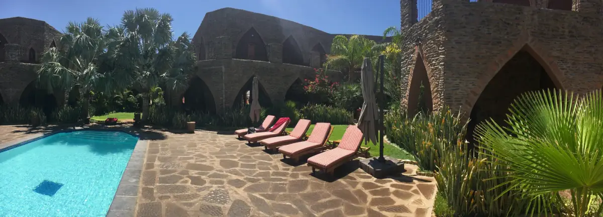 Türkises Swimmingpool mit roten Liegestühlen vor historischem Steingebäude mit Palmen und grüner Gartenanlage in der Wüste