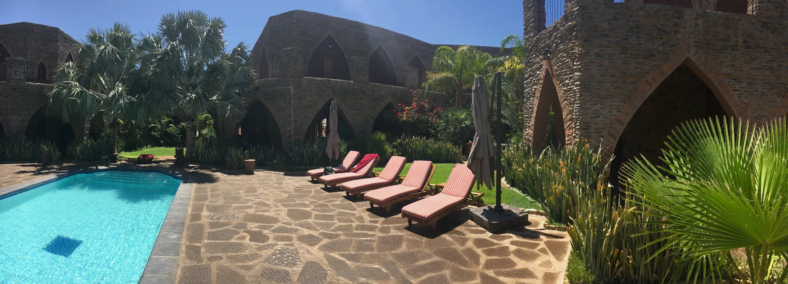 Türkises Swimmingpool mit roten Liegestühlen vor historischem Steingebäude mit Palmen und grüner Gartenanlage in der Wüste