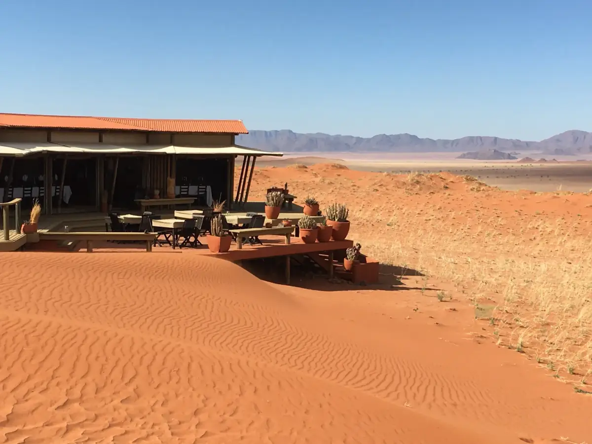 Luxuslodge mit roter Wellblechdachkonstruktion auf rotem Wüstensand, davor Sitzbereiche mit Kakteen